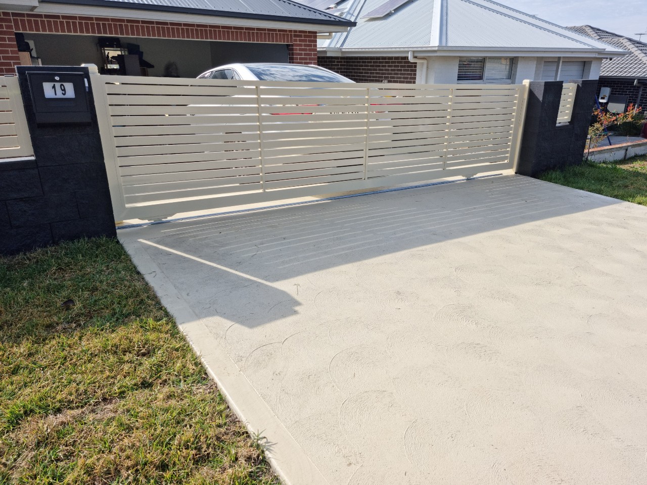 The image features a modern sliding gate installed at the front of a residential property. The gate has a slatted design painted in a light colour, providing both security and privacy while allowing visibility. The driveway is made of a smooth, light-coloured surface, and the surrounding area is well-maintained, with a small patch of grass next to the driveway. In the background, a car is partially visible, with a traditional style home and a tiled roof.